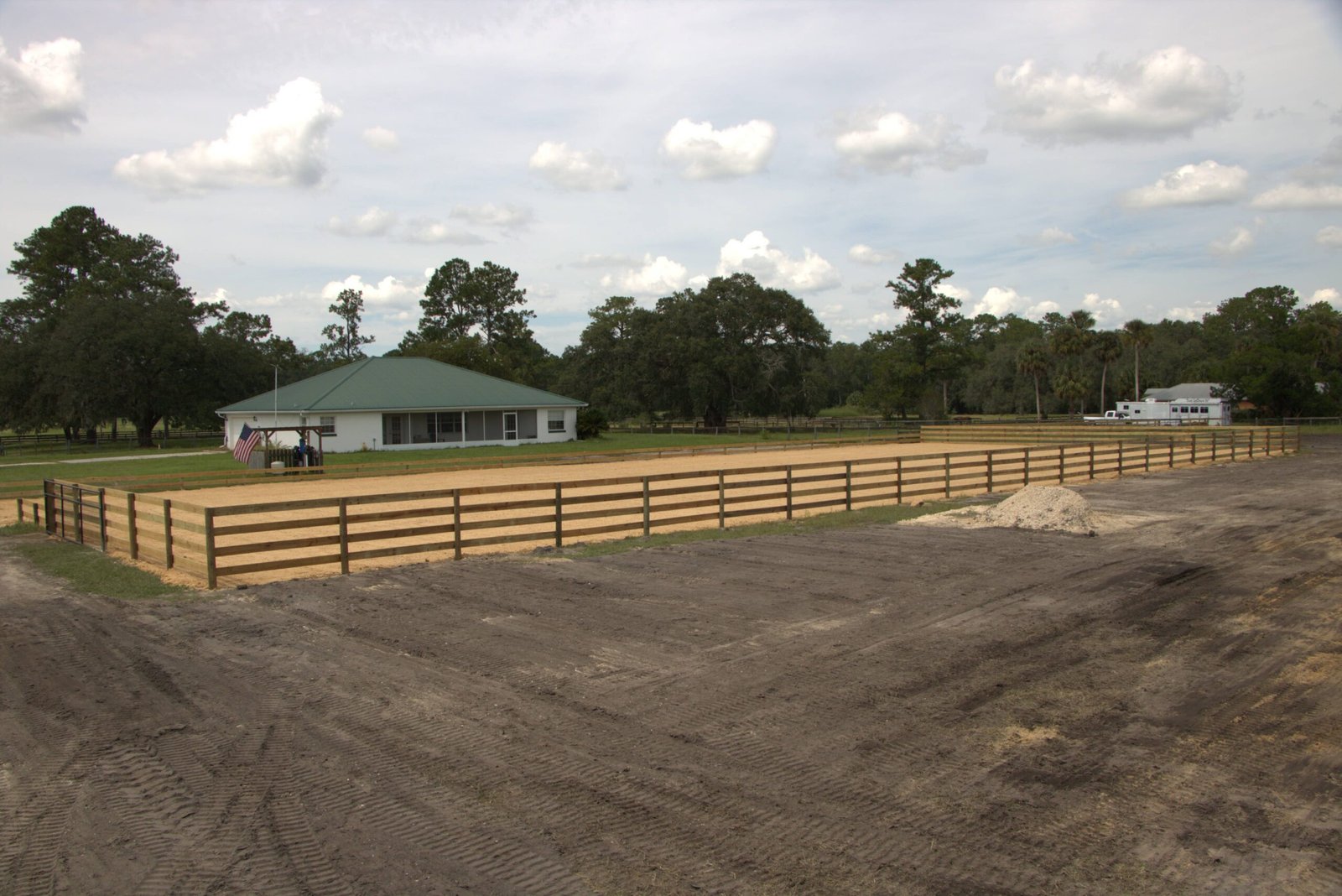 Horse arena with wooden fencing, sandy ground, and a house with a green roof in North Central Florida, showcasing suitable fencing solutions for local climate challenges.