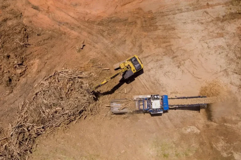 Overhead view of a yellow excavator loading a large pile of brush and wood debris into a blue and white industrial wood chipper, processing the material on a dirt site.