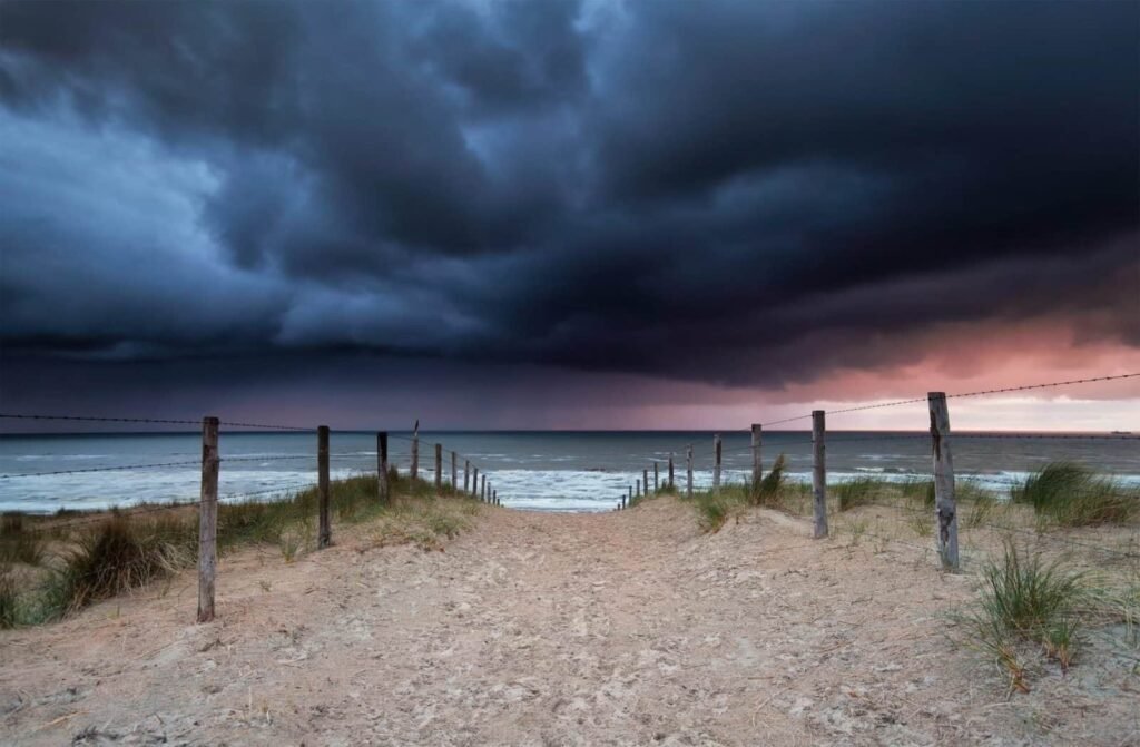 Sandy path leading down through beach dunes toward the turbulent ocean, lined by wooden posts and barbed wire, beneath a highly dramatic sky filled with heavy, dark grey and purple storm clouds.