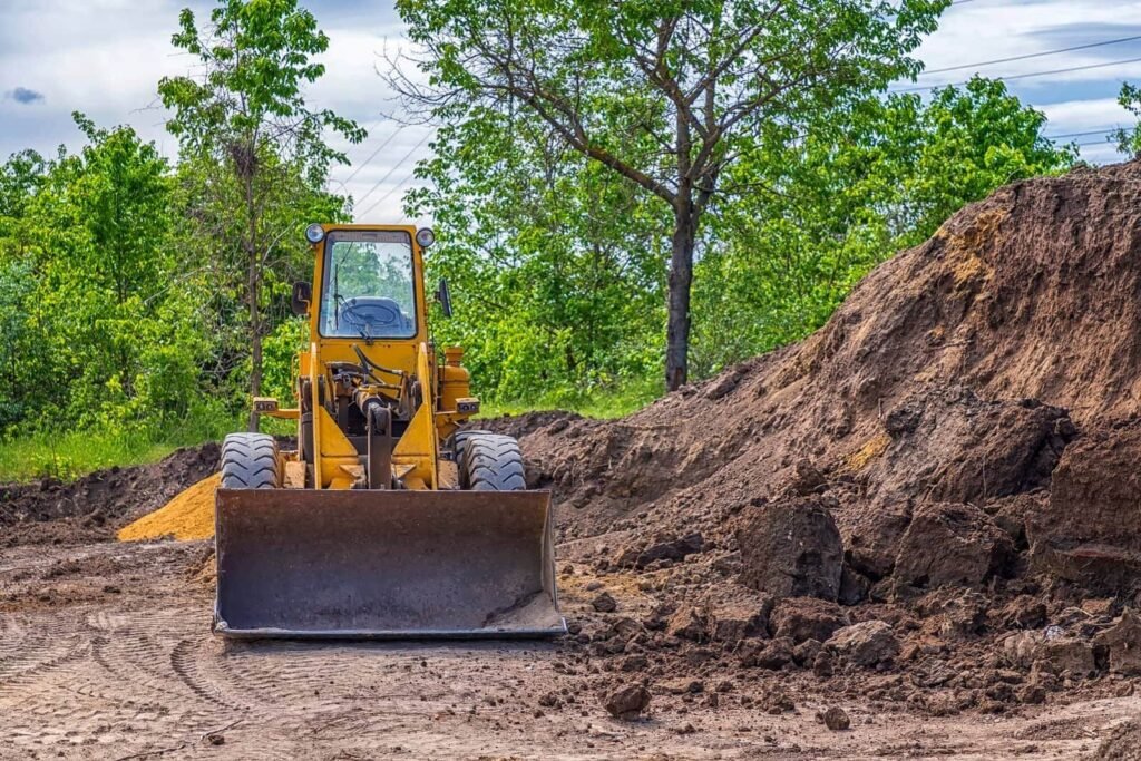 A bright yellow front loader or bulldozer with a large metal shovel blade parked on a dirt construction site. The machinery faces forward, backed by a large mound of brown earth and green trees under a cloudy sky.