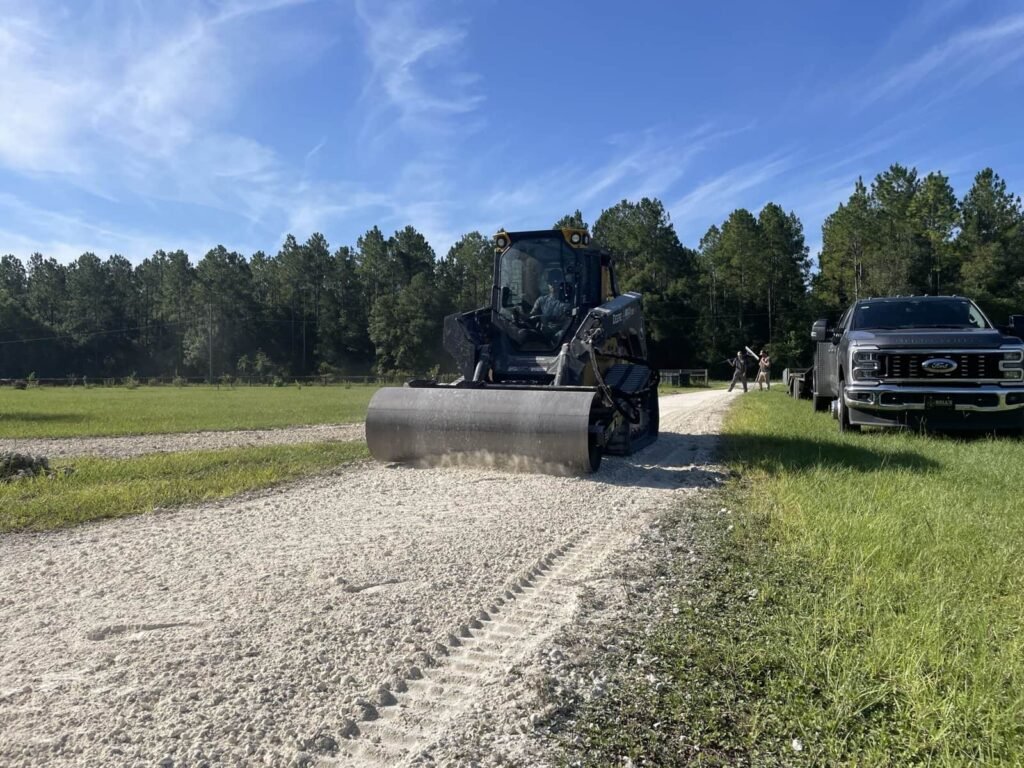 Heavy equipment compacting gravel road surface for driveway restoration, surrounded by greenery and parked trucks in North Central Florida.