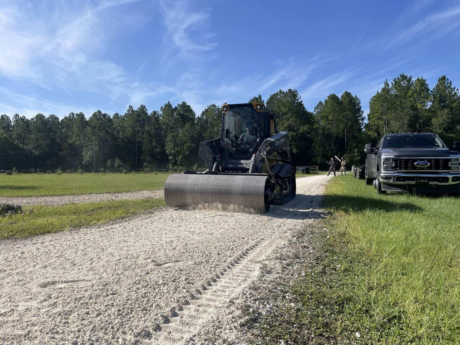 Heavy equipment compacting gravel driveway surface, surrounded by greenery and parked vehicles, illustrating professional driveway restoration services by Bell&rsquo;s Outdoor Services.