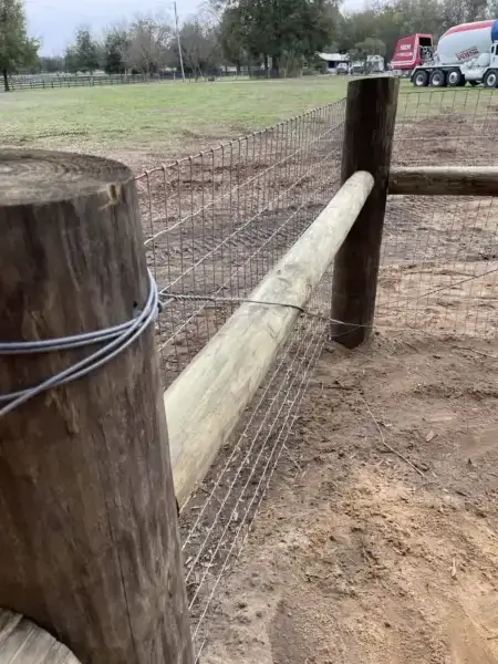Newly constructed farm fence featuring wooden posts and top rails combined with a wire mesh grid, installed in a flat green and dirt field under sunlight