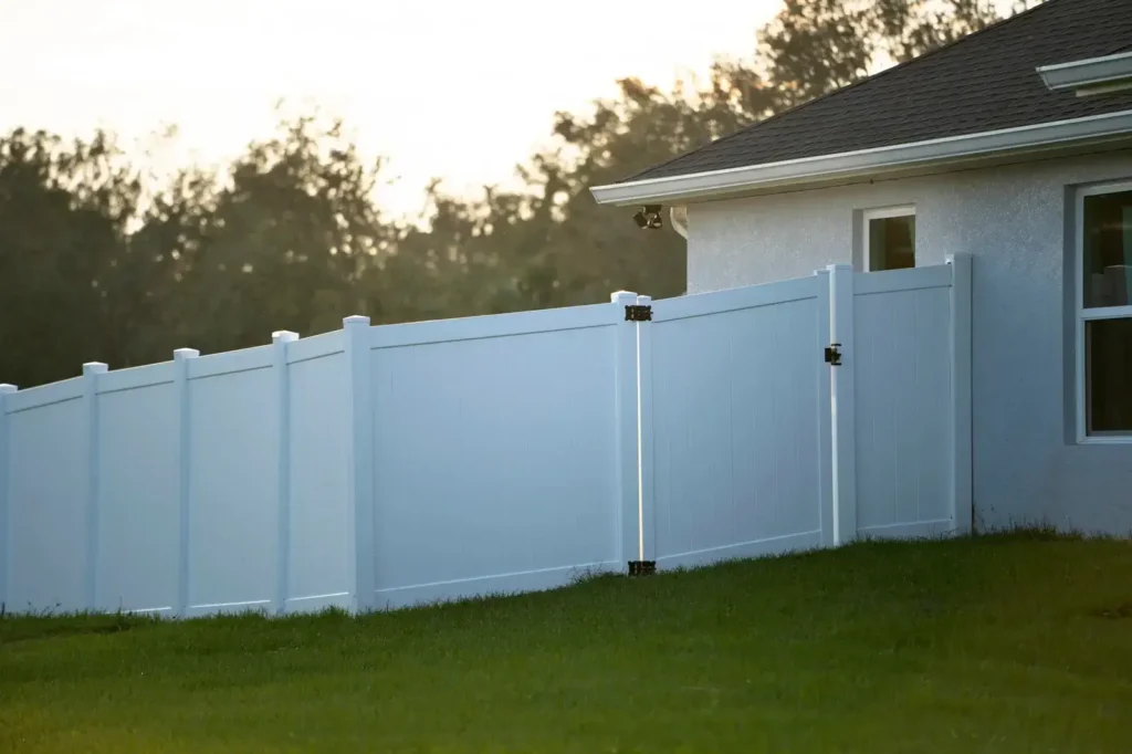 Close-up of a new, clean white vinyl privacy fence and a closed gate section on a green lawn, adjacent to the stucco exterior of a house in soft, evening light.