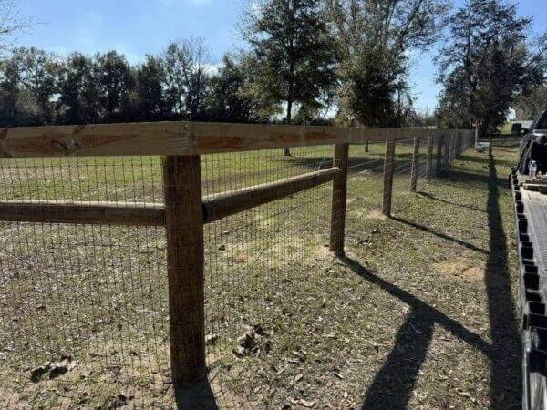 Newly constructed farm fence featuring wooden posts and top rails combined with a wire mesh grid, installed in a flat green and dirt field under sunlight.
