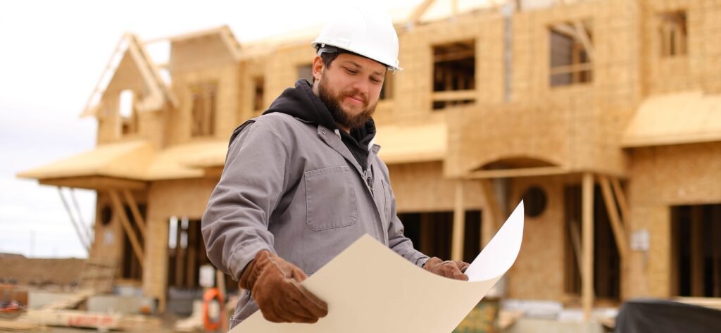 A bearded male construction worker wearing a white hard hat and work gloves reviewing architectural blueprints in front of a framed wooden house under construction.