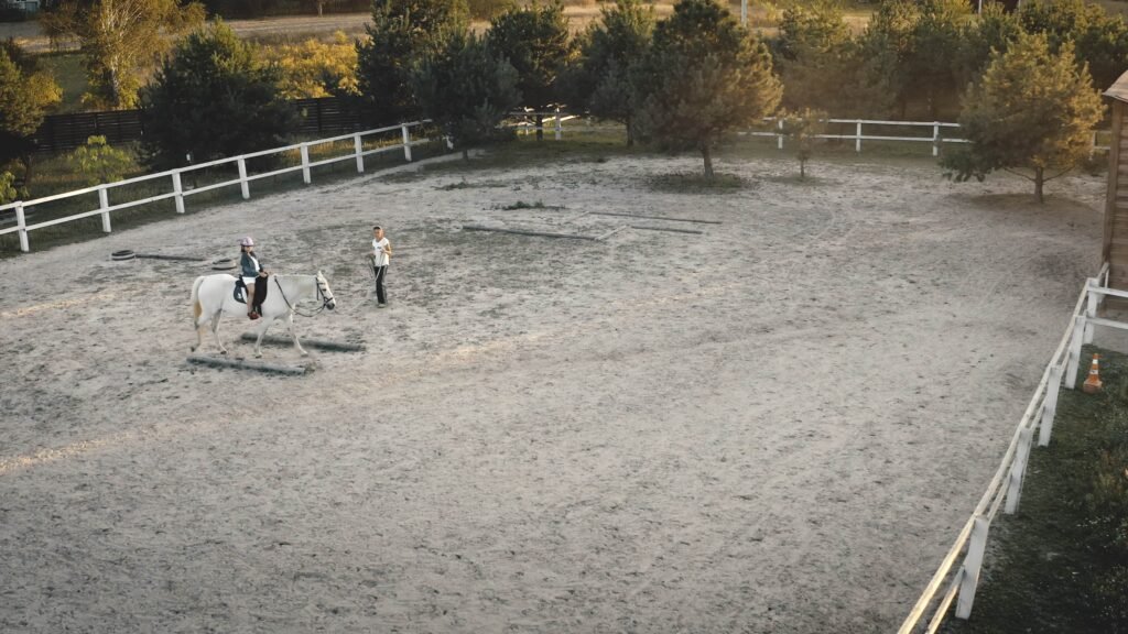 Aerial view of a child riding a white horse in an equestrian arena, with an instructor guiding them, surrounded by trees and a white fence, emphasizing equestrian training in a Florida setting.