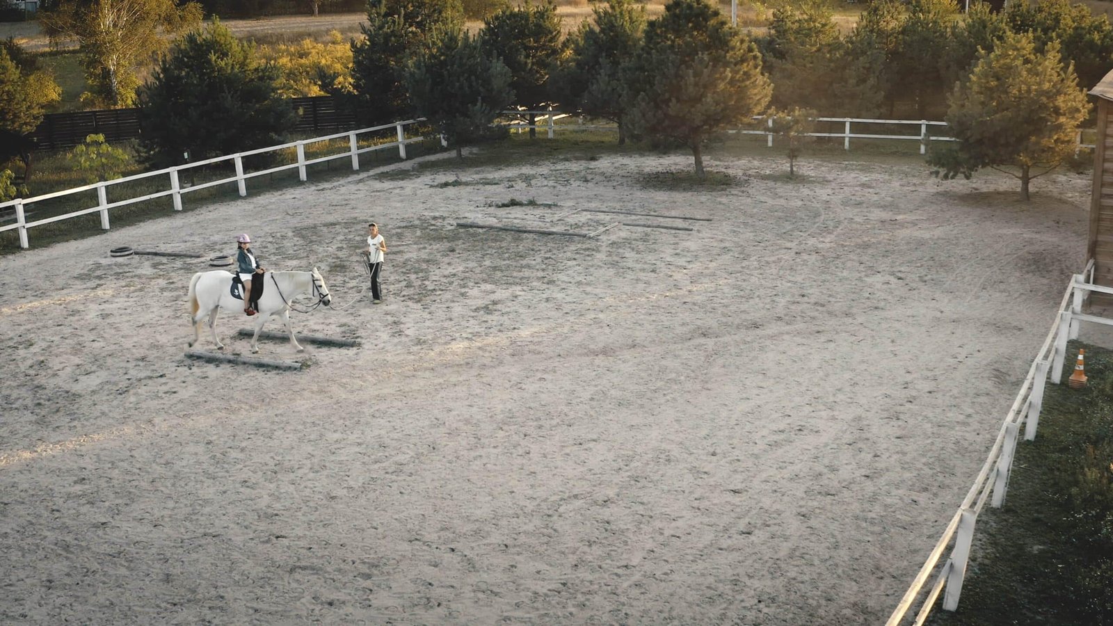 An aerial view of a young girl wearing a helmet riding a white horse over ground poles in a sandy training arena, guided by an instructor on foot.