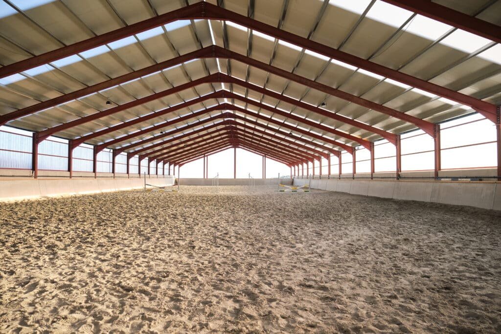 Interior view of a vast, empty indoor horse riding hall featuring a red steel frame, translucent roof panels for natural light, and professional sand footing for equestrian training.