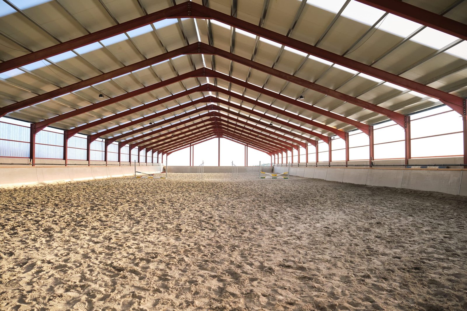 Interior view of a vast, empty indoor horse riding hall featuring a red steel frame, translucent roof panels for natural light, and professional sand footing for equestrian training.